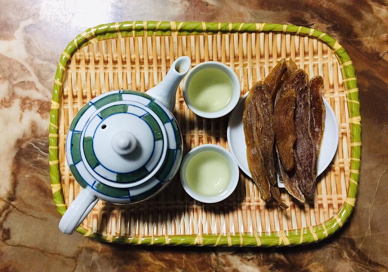 Đĩa khoai deo Quảng Bình màu nâu cánh gián đặt cạnh ấm trà và hai chén nước chè xanh trên khay tre